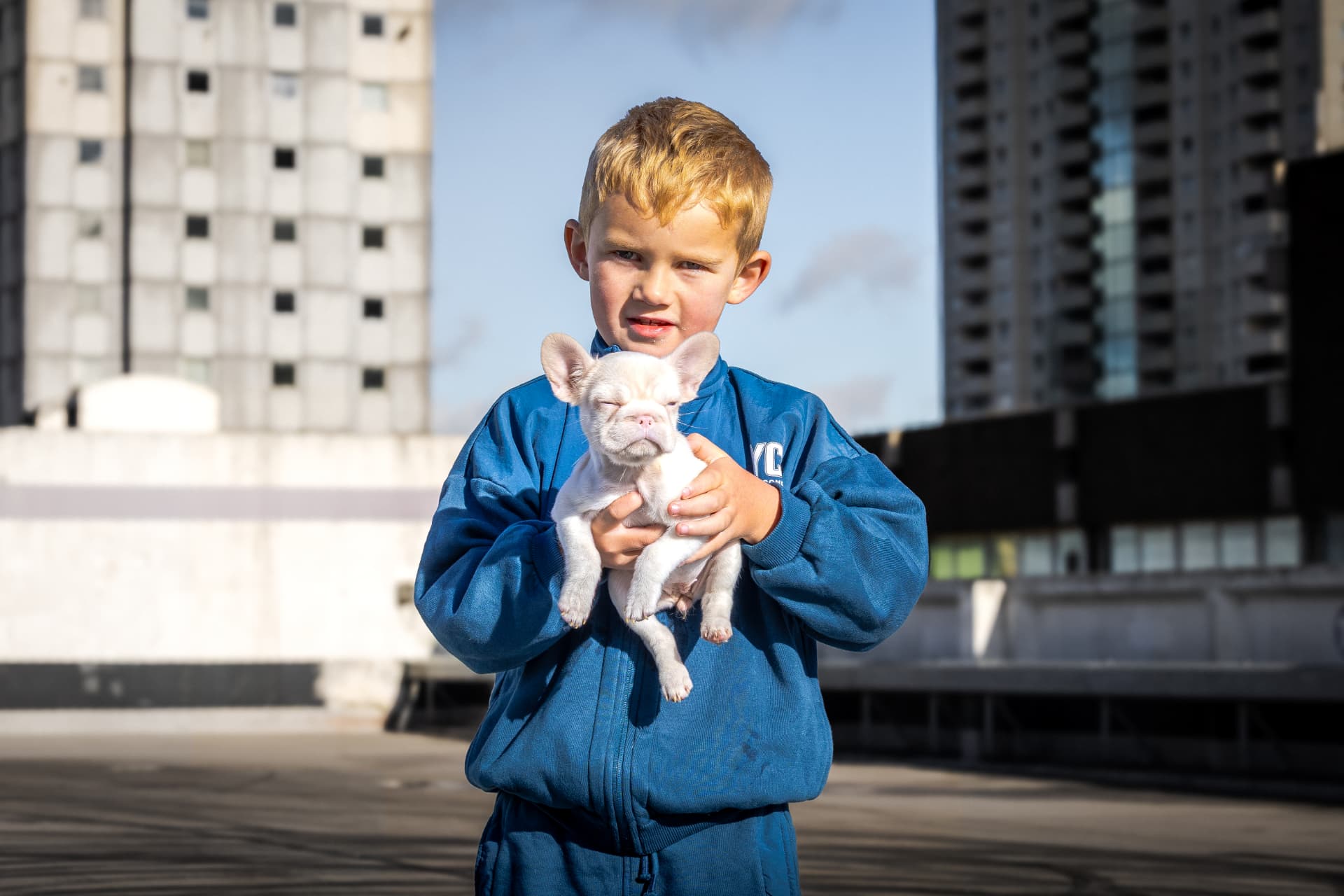 Young boy in a blue tracksuit holding a cream French Bulldog puppy on a London rooftop